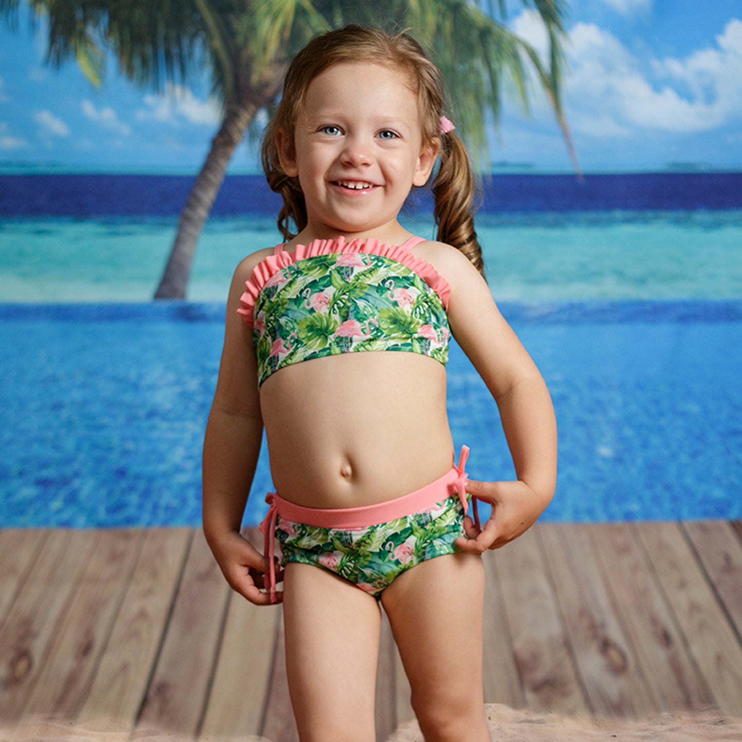 Modellen fotózott kétrészes kislány bikini szett, rózsaszín flamingós mintával, színnel azonos vállpántokkal és fodorral, elölről fotózva tengerparti háttér előtt.