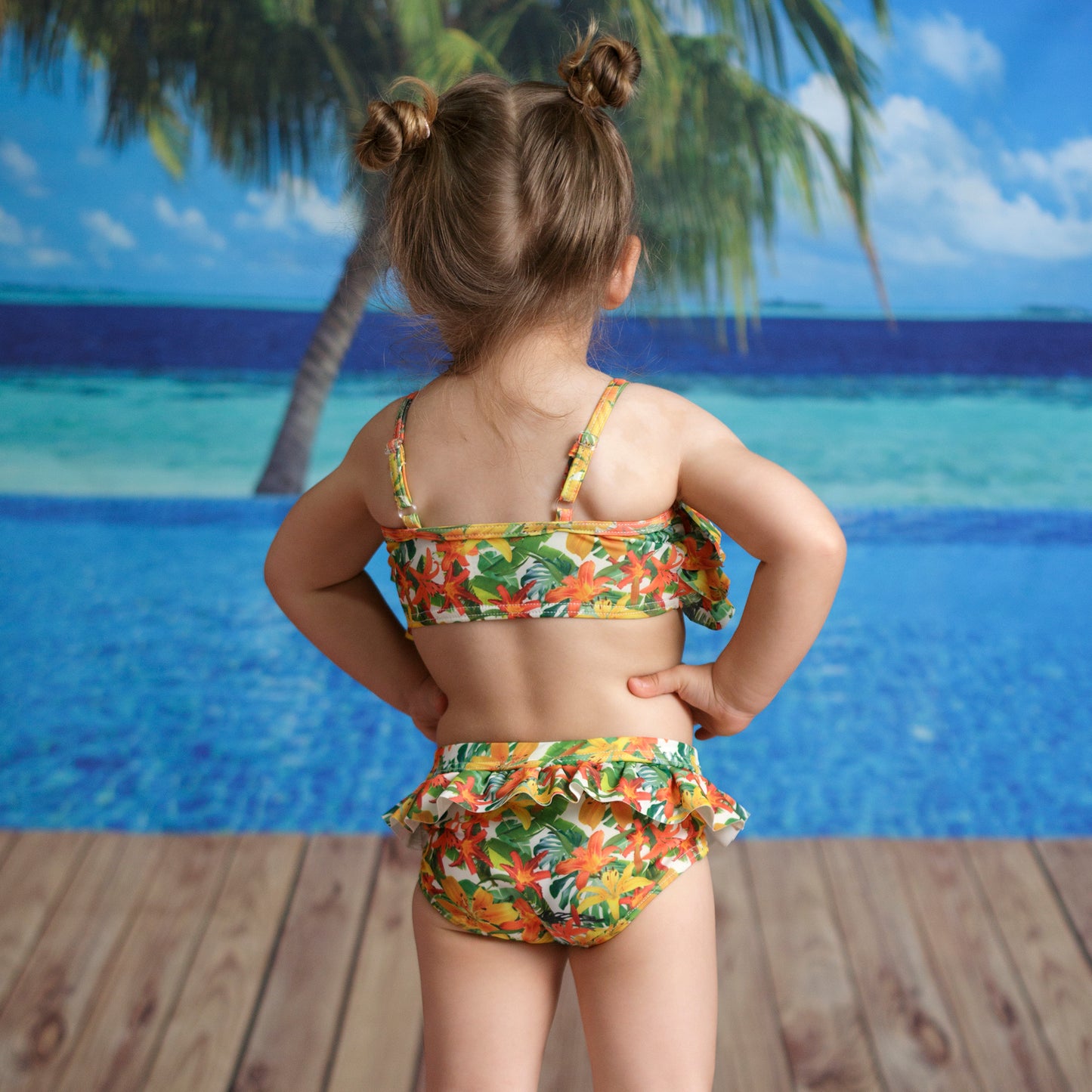 Hawaii virágos kislány kétrészes bikini szett, hátulról fotózva modellen, állítható vállpántokkal, fodros klasszikus szabású alsóval, tengerparti háttér előtt.
