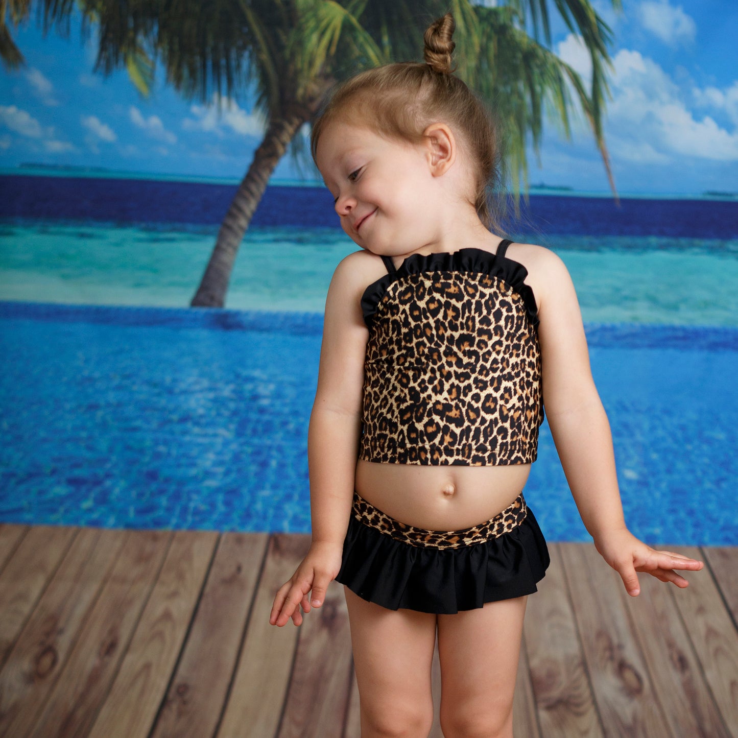 Leopárd mintás kislány bikini szett, modellen fotózva, fodros és vállpántos tankini felsővel, klasszikus szabású fodros alsóval, tengerparti háttérrel előről.
