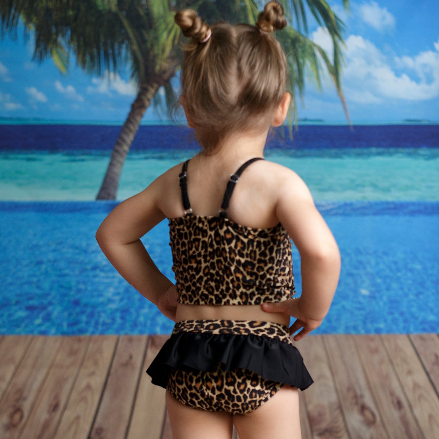 Leopárd mintás, kétrészes kislány bikini szett modellen hátulról fotózva, tengerparti háttér előtt, állítható vállpántokkal és fodros alsóval.