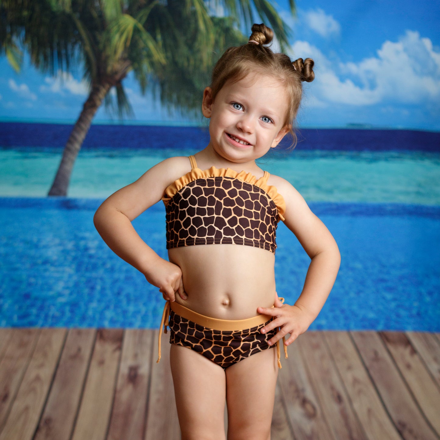 Zsiráf mintás kislány kétrészes bikini szett,  mellkason vékony fodorral díszített vállpántos felsővel és gumírozott oldalt díszmegkötős alsóval, modellen fotózva tengerparti háttér előtt.